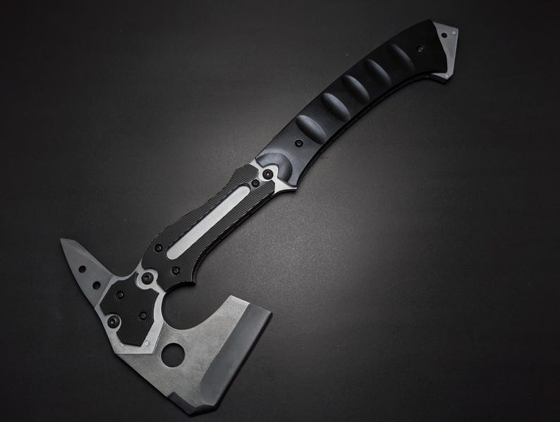 May include: A tactical hatchet featuring a matte black curved handle with textured grip sections and a silver metal inlay. The head is constructed from dark grey metal, showcasing a sharp blade edge, a pointed spike, and a circular cutout.