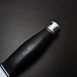 May include: Close-up of a knife handle. The handle is black with a textured surface and a silver bolster and end cap. A thin white line separates the blade from the handle.