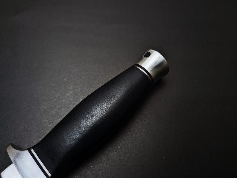 May include: Close-up of a knife handle. The handle is black with a textured surface and a silver bolster and end cap. A thin white line separates the blade from the handle.
