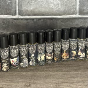 May include: A collection of thirteen cylindrical glass roller bottles with black caps, each featuring a label with the brand name Covet and unique illustrations including a chalice, a mask, a skull, a clock tower, books, and an axe.