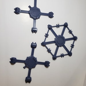 Op de afbeelding: Drie grijze plastic modelbases voor miniatuurfiguren. Twee bases hebben een ontwerp met vier armen en één basis heeft een ontwerp met zes armen.
