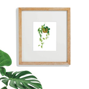 Puede incluir: Una impresión artística enmarcada que presenta una planta colgante en una maceta marrón. La planta tiene hojas verdes y enredaderas colgantes. La obra de arte está sobre un fondo blanco y enmarcada en un marco de madera clara. Una gran hoja verde está en la esquina inferior izquierda.