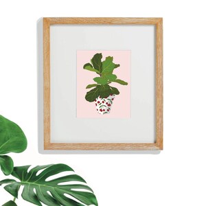 Op de afbeelding: Een ingelijste print van een groene vioolbladficus in een witte pot met een rood kersenpatroon op een lichtroze achtergrond.