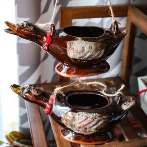 May include: Two brown ceramic duck planters hanging from twine. The ducks have red accents and a white glaze.