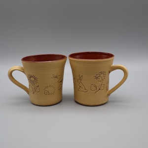May include: Two ceramic mugs with a light tan exterior and a dark reddish-brown interior. Each mug features a handle and is decorated with etched line drawings of sunflowers, a hedgehog, a rabbit, and small leaves around the outer surface.