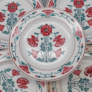 Könnte beinhalten: Eine Sammlung weißer Keramikteller mit einem rot-grünen Blumenmuster. Jeder Teller hat ein zentrales Mohnblumenmotiv und einen dekorativen Rand. Geeignet zum Servieren von Mahlzeiten.
