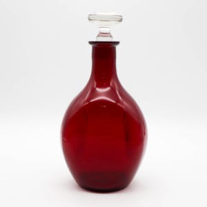May include: A deep red glass decanter with a clear glass stopper. The decanter has a rounded body and a long neck. The stopper is a flat, circular shape. The decanter is likely used for serving wine or other beverages.