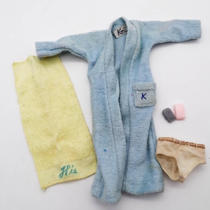 Pode incluir: Um roupão de banho azul claro com um bolso com um patch azul "K", uma toalha amarela bordada com "His", um par de roupa interior bege e duas pequenas barras de sabão. O roupão tem uma etiqueta que diz "Ken".
