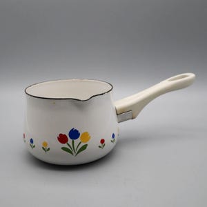 May include: A white enamel saucepan featuring a floral pattern of red, blue, and yellow tulips with green stems. The pot has a dark rim, a small pouring spout, and a cream-colored handle attached with a silver-toned metal connector.