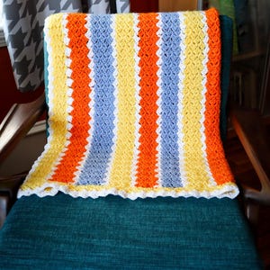May include: A colorful, handmade crochet blanket with vertical stripes of orange, yellow, and blue. The blanket has a white scalloped edge and is draped over a teal chair. The background includes a window with patterned curtains.