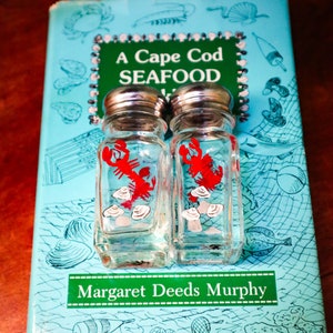Könnte beinhalten: Ein Vintage-Kochbuch mit dem Titel "A Cape Cod Seafood Cookbook" von Margaret Deeds Murphy, mit einem Paar Salz- und Pfefferstreuern mit einem Hummer- und Muschel-Design.