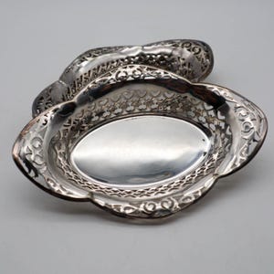 Può includere: Un piatto da portata ovale color argento con intricati dettagli traforati. Il piatto ha un interno liscio e riflettente e un bordo decorativo smerlato. Un pezzo più piccolo abbinato è posizionato dietro.