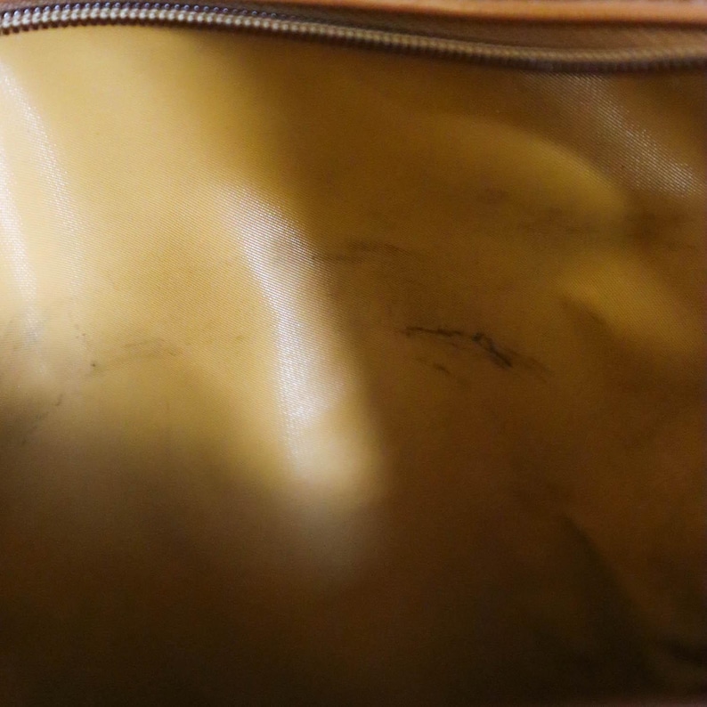 Pu&ograve; includere: Primo piano dell'interno di una borsa. La fodera &egrave; di un colore oro chiaro con una lucentezza satinata. Sono visibili macchie scure sul tessuto. La cerniera &egrave; visibile nella parte superiore dell'immagine.