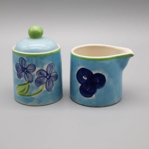May include: A ceramic sugar bowl with a lid and a creamer. Both are light blue with hand-painted dark blue floral designs and green accents. The sugar bowl has a green knob on the lid.