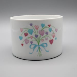 May include: White ceramic planter with a colorful heart bouquet design. The bouquet features hearts in shades of pink, blue, and purple, tied with a light blue ribbon. The planter is cylindrical and has a smooth, glossy finish.