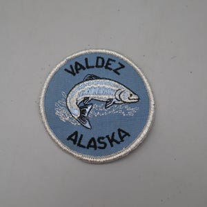 May include: A circular embroidered patch with a light blue background and a white stitched border. The patch features a silver and white fish graphic in the center, with the black text VALDEZ arched above and ALASKA arched below the fish.
