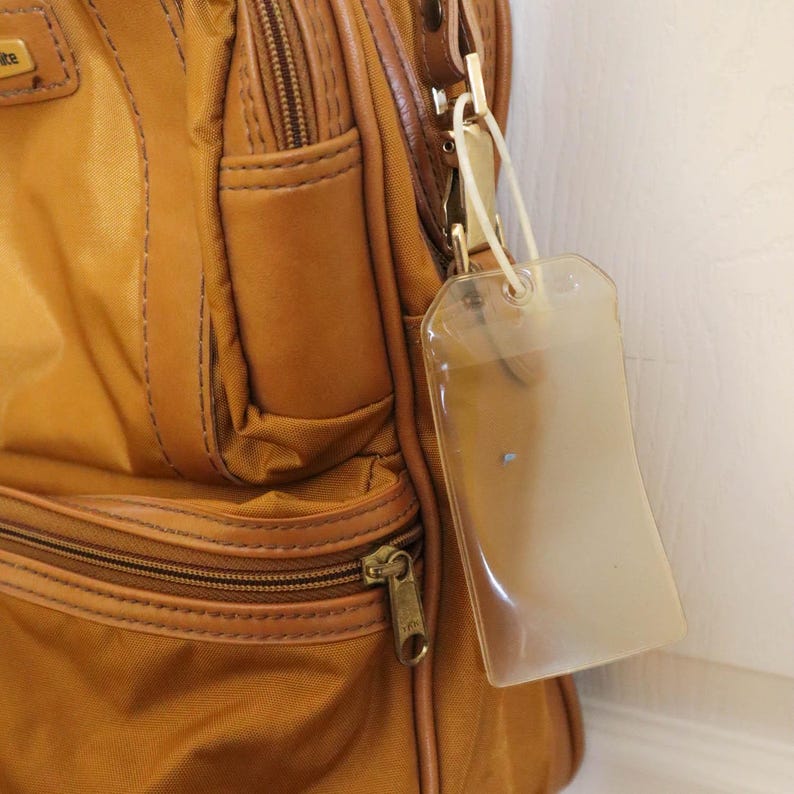 Pu&ograve; includere: Primo piano di una borsa da viaggio beige con un'etichetta per bagagli trasparente attaccata. La borsa ha una tasca con cerniera, finiture in pelle marrone e una cerniera color oro. L'etichetta per bagagli &egrave; rettangolare.