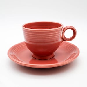 May include: A coral-colored teacup and saucer set. The cup has a rounded shape with a handle and horizontal ridged detailing. The saucer is round and matches the cup's color and ridged design. The set is made of ceramic.