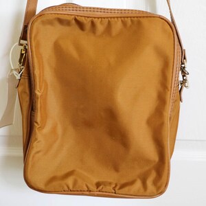 Pu&ograve; includere: Una borsa a tracolla rettangolare color senape con cinturino e finiture in pelle marrone. La borsa ha un esterno in nylon strutturato e una chiusura in metallo. Un'etichetta vuota pende dalla tracolla.