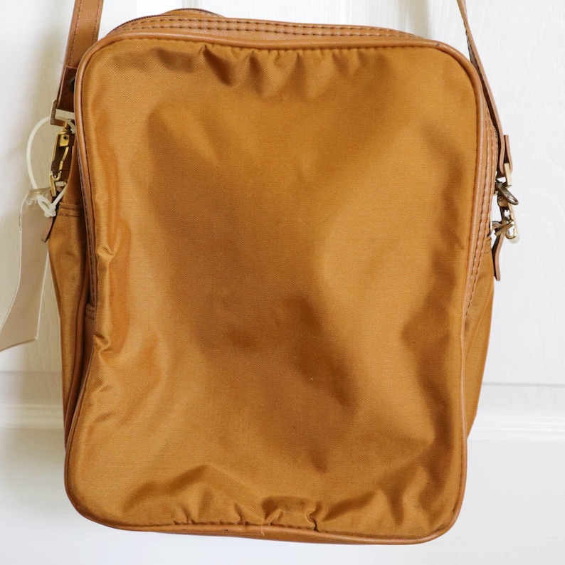 Pu&ograve; includere: Una borsa a tracolla rettangolare color senape con cinturino e finiture in pelle marrone. La borsa ha un esterno in nylon strutturato e una chiusura in metallo. Un'etichetta vuota pende dalla tracolla.