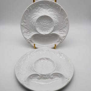 May include: Two white ceramic serving dishes with floral and leaf relief patterns. Each dish has a central circular well and a curved, elongated indentation. The dishes are displayed on gold-colored stands.