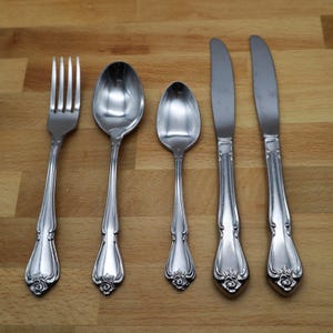 Pode incluir: Um conjunto de cinco utensílios de cor prateada: um garfo, duas colheres e duas facas. Os utensílios têm detalhes florais decorativos na base das pegas. Os itens estão dispostos sobre uma superfície de madeira castanha clara.