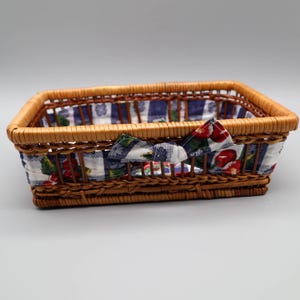 May include: Rectangular wicker basket with a blue and white checkered fabric lining and a decorative bow. The fabric features a pattern of red berries and green leaves. The basket is made of woven brown wicker.