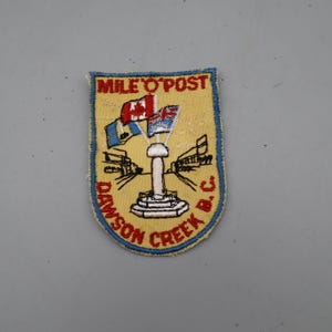 May include: A rectangular embroidered patch with a light yellow background and a blue border. It features a central white monument with two flags, red text reading MILE O'POST at the top, and red text reading DAWSON CREEK B.C. at the bottom.