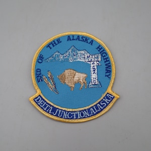 May include: A circular embroidered patch with a light blue background and a yellow border. It features a brown bison, a white mountain range, and a white signpost reading MILE 1422. Text reads END OF THE ALASKA HIGHWAY and DELTA JUNCTION ALASKA.