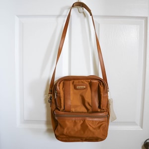 Pu&ograve; includere: Una borsa a tracolla Samsonite marrone con una lunga tracolla regolabile. La borsa ha una forma rettangolare con una tasca frontale con cerniera e un piccolo logo. La borsa &egrave; appesa a una porta bianca.