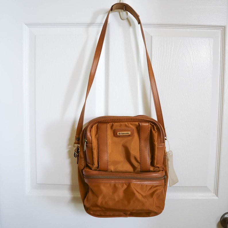 Pu&ograve; includere: Una borsa a tracolla Samsonite marrone con una lunga tracolla regolabile. La borsa ha una forma rettangolare con una tasca frontale con cerniera e un piccolo logo. La borsa &egrave; appesa a una porta bianca.