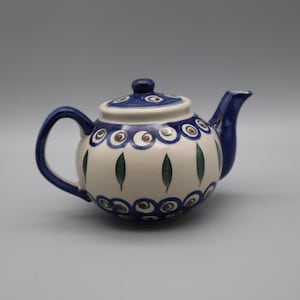 May include: A ceramic teapot with a cream-coloured base, featuring a blue handle, spout, and lid. The body is decorated with rows of blue and brown circular patterns at the top and bottom, with vertical green leaf-like motifs around the centre.