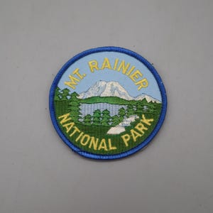 May include: A circular embroidered patch featuring a landscape of Mt. Rainier National Park. The design includes a snow-capped mountain, a blue lake, and green pine trees. The text MT. RAINIER NATIONAL PARK is stitched in yellow thread.