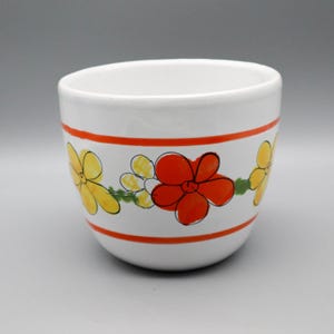 以下が含まれることがあります： 鮮やかな赤と黄色の花が手描きされた水平な帯模様が特徴の白い陶器製のボウル。丸みを帯びた容器の外側には、花柄を縁取る細いオレンジ色の線が描かれており、その模様が容器の周囲をぐるりと囲んでいます。