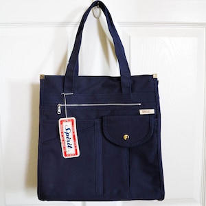 May include: Navy blue tote bag with a rectangular shape, featuring a zipper closure and multiple pockets. The bag has a tag that reads "Spirit" in a red and white design. The bag is hanging on a white door.