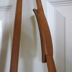 Pu&ograve; includere: Primo piano di una tracolla in pelle marrone con una sezione imbottita per il comfort. Il cinturino ha cuciture visibili lungo i bordi e sembra essere attaccato a una borsa. Lo sfondo &egrave; una porta bianca.