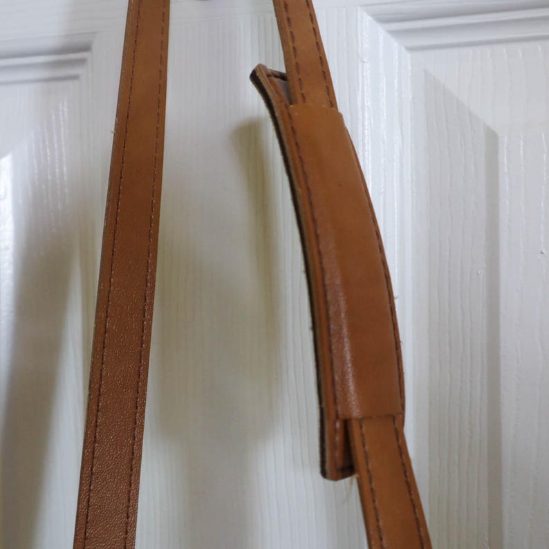 Pu&ograve; includere: Primo piano di una tracolla in pelle marrone con una sezione imbottita per il comfort. Il cinturino ha cuciture visibili lungo i bordi e sembra essere attaccato a una borsa. Lo sfondo &egrave; una porta bianca.