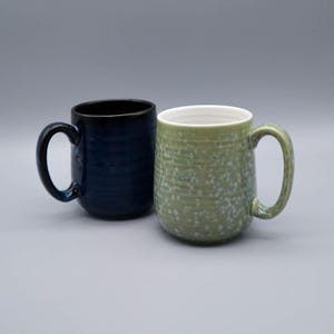 Puede incluir: Dos tazas de cerámica de forma cilíndrica con un asa lateral. Una taza presenta un esmalte azul marino profundo con estrías horizontales, mientras que la otra tiene un esmalte verde salvia claro con una textura moteada y estrías horizontales.