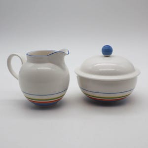 May include: A white ceramic creamer and sugar bowl set. The creamer has a handle and spout, with blue trim. The sugar bowl has a lid with a blue knob. Both pieces have colorful stripes around the base.
