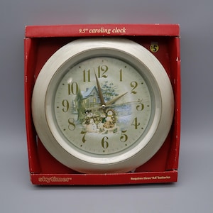 May include: A 9.5-inch round wall clock with a white frame, housed in a red cardboard box. The clock face features a winter scene with snowmen and a house, gold-colored numbers, and gold hands. The text Skytimer is printed on the box.