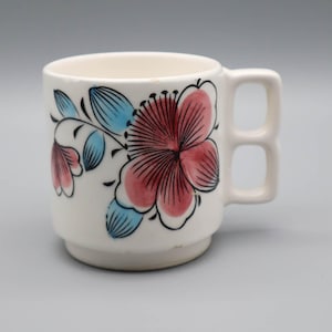 May include: White ceramic mug with a floral design. The flowers are pink with black outlines and the leaves are blue. The mug has a square handle and a stepped base.