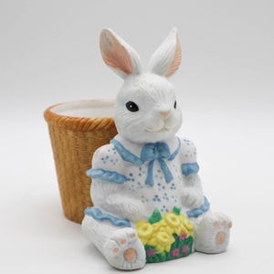 Puede incluir: Figura de conejo de cerámica blanca con vestido ribeteado en azul y flores amarillas. Una pequeña cesta de mimbre está al lado. El conejo tiene detalles rosas en las orejas y patas. Decoración de Pascua.