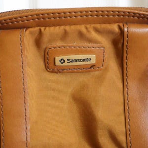 Pu&ograve; includere: Primo piano di una valigia Samsonite vintage. La borsa presenta un pannello centrale in nylon color cachi con finiture in pelle marrone e una chiusura con cerniera. Il logo Samsonite &egrave; visualizzato su una toppa rettangolare in pelle.