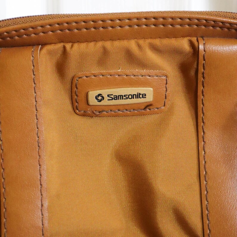 Pu&ograve; includere: Primo piano di una valigia Samsonite vintage. La borsa presenta un pannello centrale in nylon color cachi con finiture in pelle marrone e una chiusura con cerniera. Il logo Samsonite &egrave; visualizzato su una toppa rettangolare in pelle.
