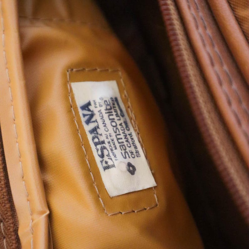 Pu&ograve; includere: Primo piano dell'interno di una borsa da viaggio Samsonite marrone. Un'etichetta rettangolare &egrave; cucita all'interno, con il testo "ESPANA" e "Samsonite of Canada Limited". L'etichetta &egrave; bianca con testo blu e un bordo marrone. L'interno della borsa &egrave; marrone chiaro.