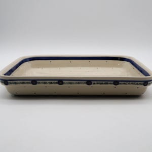 Puede incluir: Fuente rectangular de cerámica color crema con detalles azules. El borde presenta un borde azul sólido y una banda decorativa con motivos florales azules. Pequeños puntos azules están dispersos por la superficie.