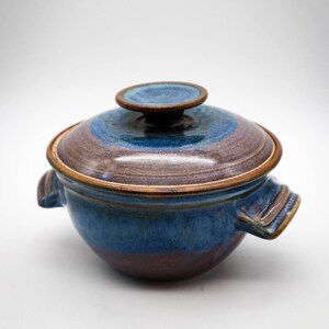 May include: A blue and brown ceramic casserole dish with a lid and two handles. The dish has a glazed finish and a rustic look.