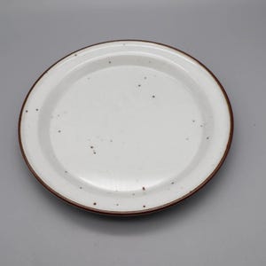 Op de afbeelding: Een witte keramische plaat met een gespikkeld ontwerp en een bruine rand. De plaat is rond en heeft een licht verhoogde rand. De plaat heeft een diameter van ongeveer 25 cm.