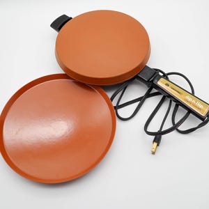 Peut inclure: Un crêpier orange Universal Dip n' Flip avec une surface de cuisson ronde et une assiette de service séparée. L'appareil a une poignée et un cordon noirs, et le texte "Dip n' Flip Crepe Maker" est visible sur la poignée.