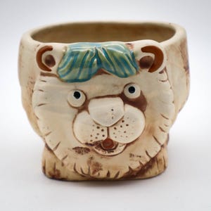 May include: A ceramic planter shaped like a lion's head with a green and blue glaze on the top. The planter has a white base with brown accents.